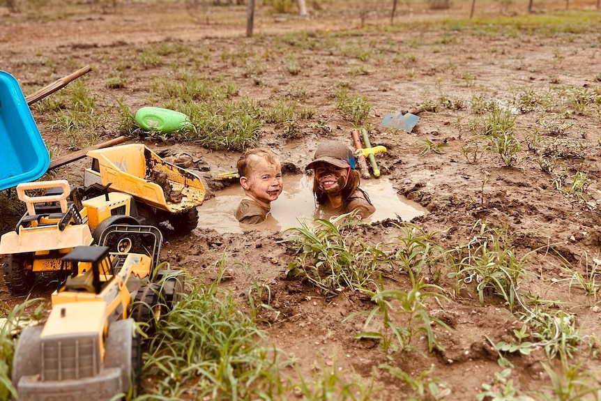 Two children are sitting in a deep hole filled with muddy water. They are smiling and have mud on their faces.