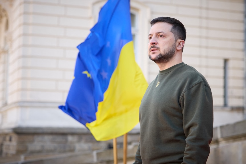 A man in army fatigues stands in front of the Ukrainian flag