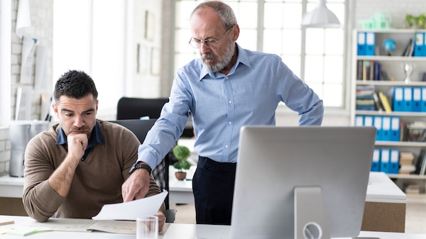 Male coworkers discussing in office, looking at documents. One man looks bored and exhausted
