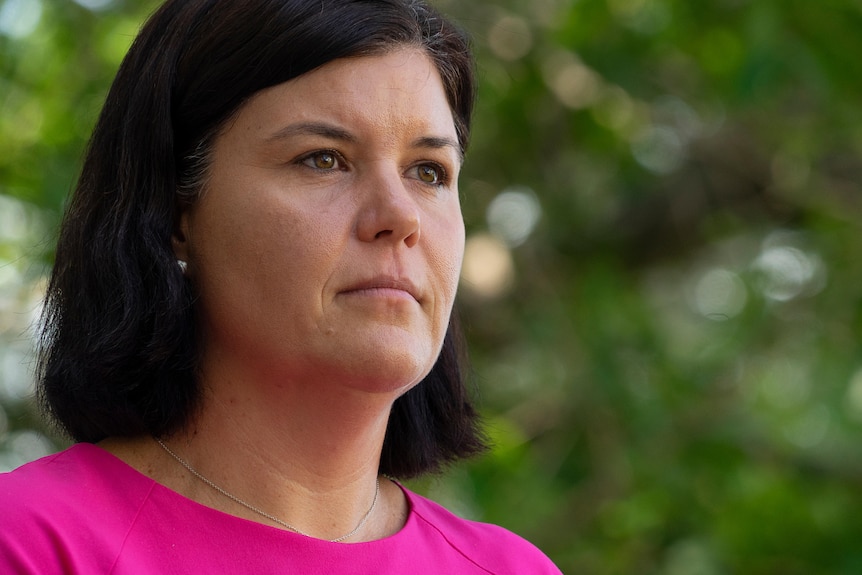 A woman with dark, shoulder-length hair is wearing a pink shirt and looking serious into the distance.