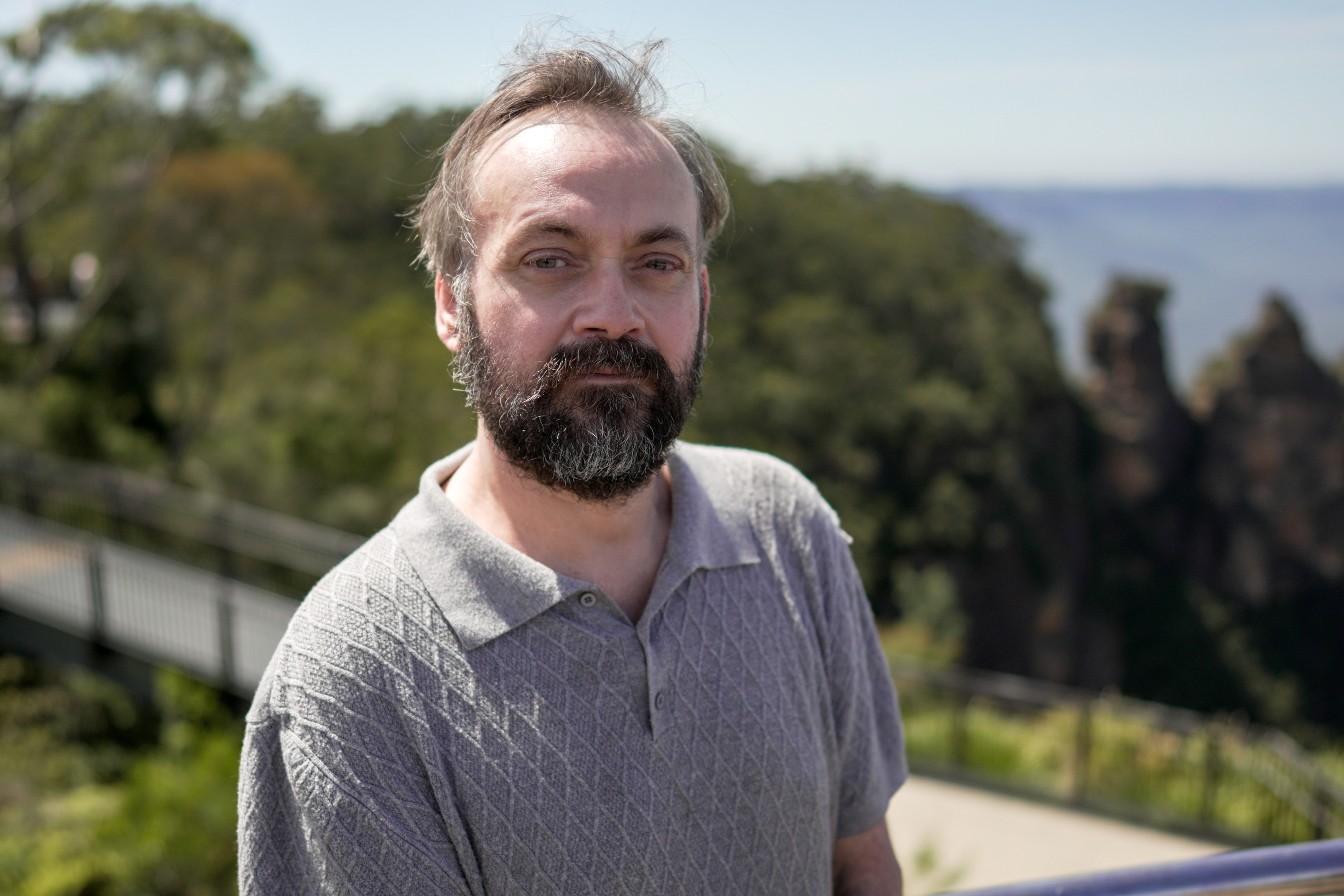 A man in a grey shirt and beard stands outside looking at camera.
