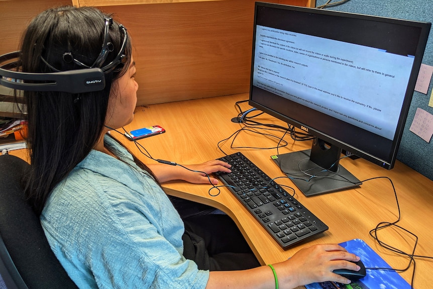 A person sits at a computer, wearing a headset with multiple probes against the head.