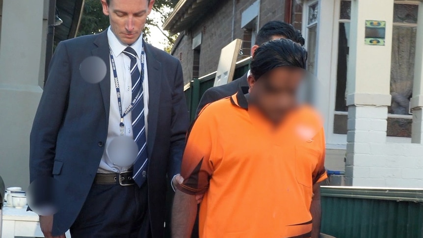 A man wearing an orange shirt walks in front of man with a suit.