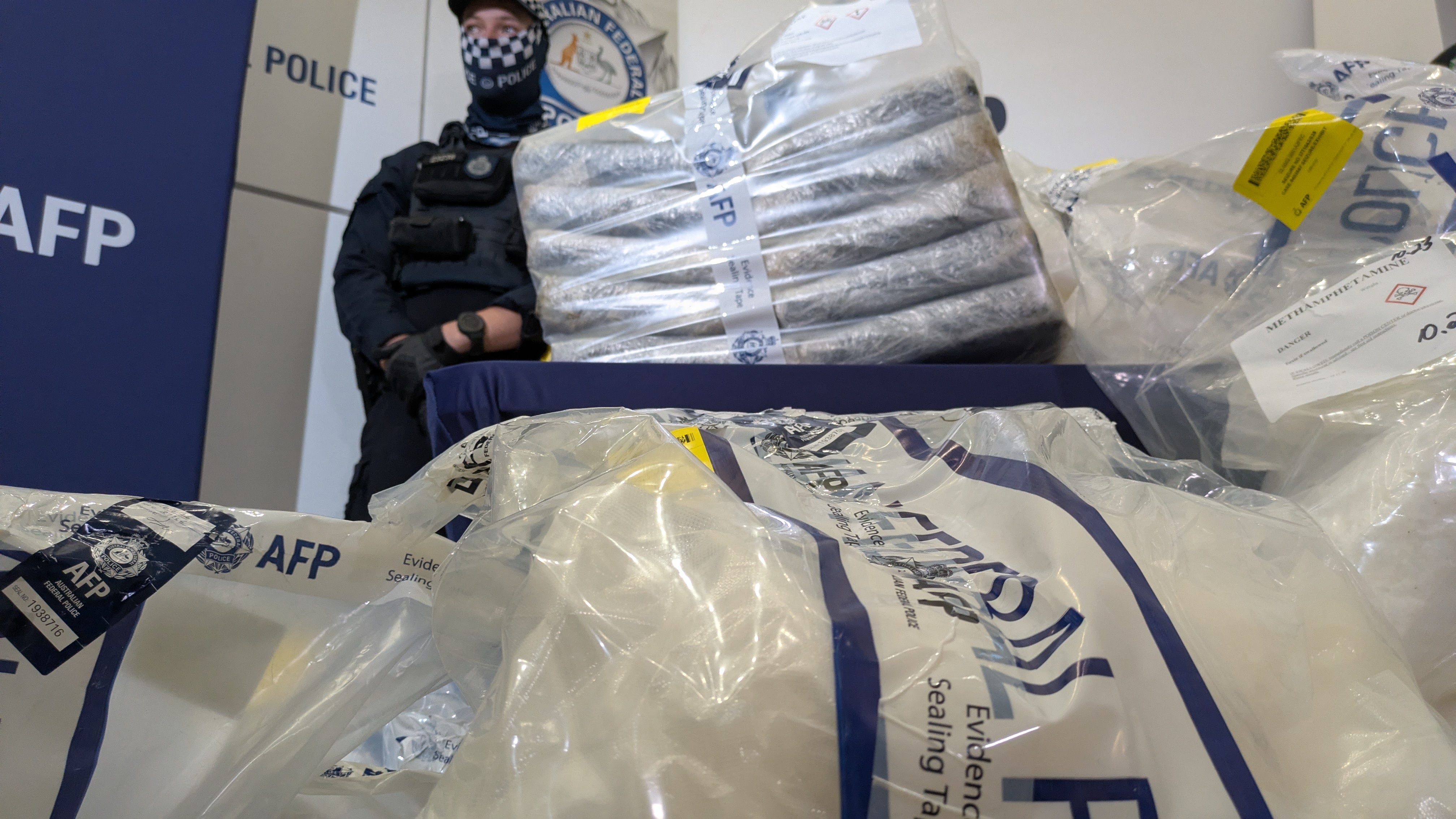 A police officer stands behind evidence bags full of drugs.