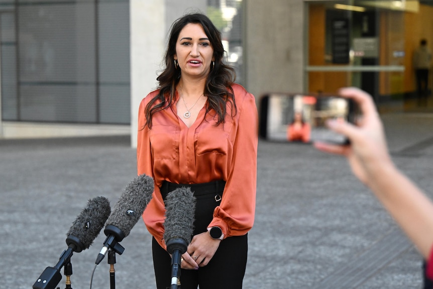 a woman wearing a silk blouse speaks into media microphones outside a courthouse