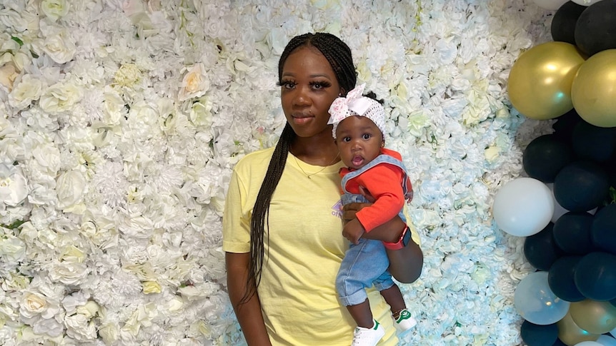 A mother holds her baby in front of a flower wall