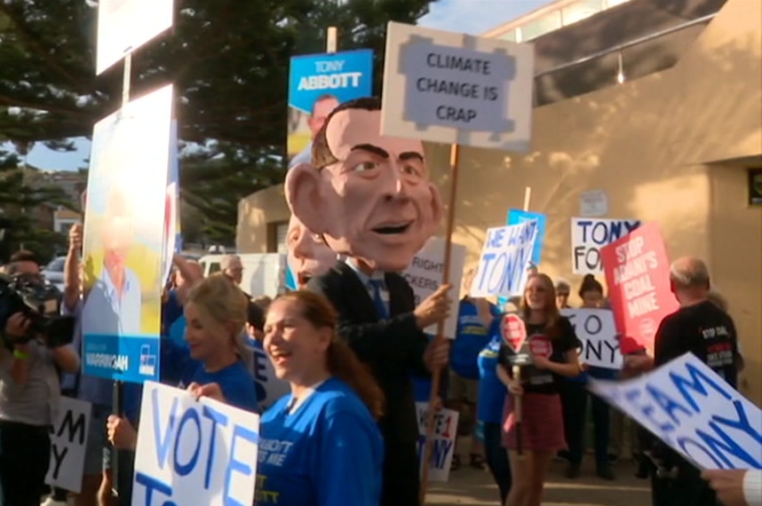 A man with a giant Tony Abbott head and holding a "climate change is crap" sign is surrounded by others with "vote Tony" signs.