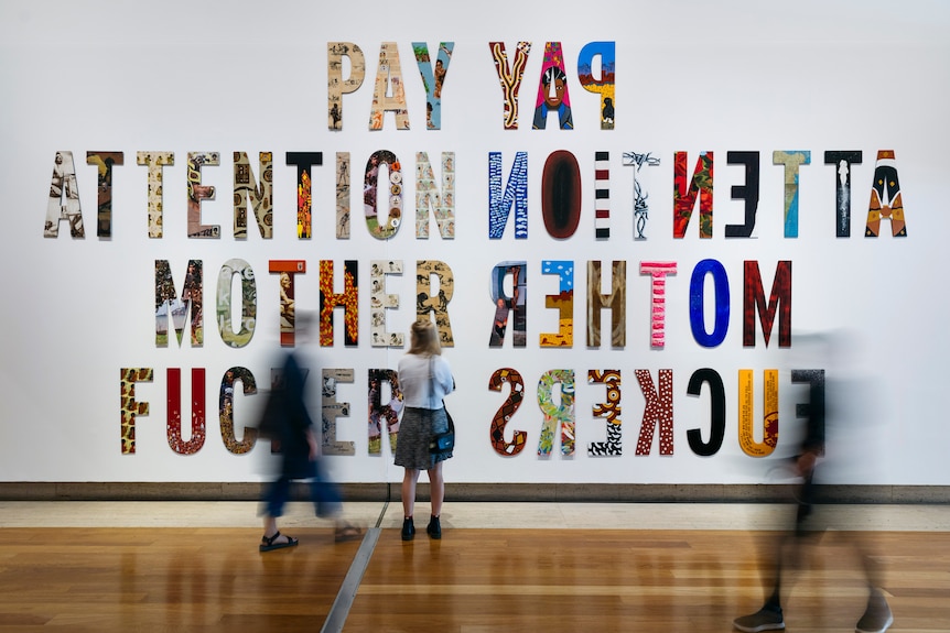 Gallery guests mill in front of the artwork which spells out ‘PAY ATTENTION MOTHERF***ERS’ in a variety of large letters.