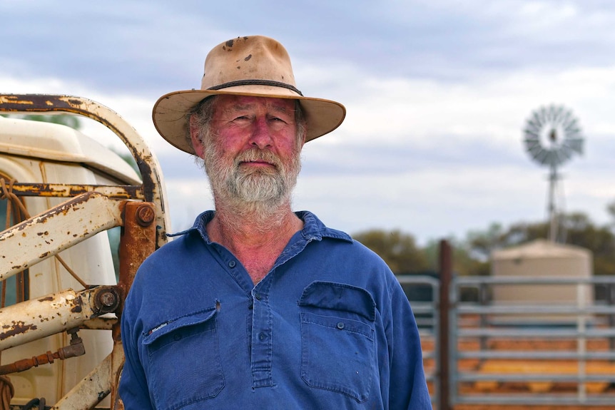 A man wearing a hat stands on a pastoral property looking at the camera