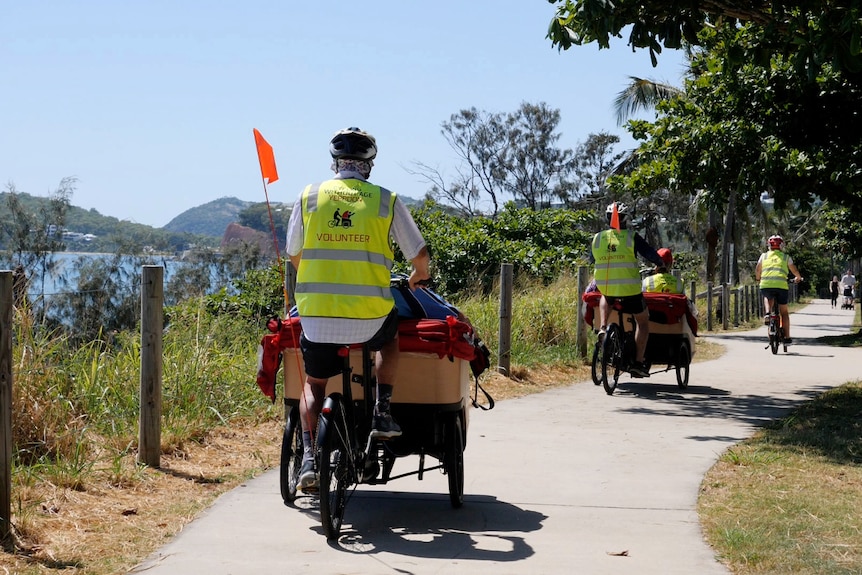 Two trishaws carrying people, pilot at the front on a bicycle, riding along a footpath next to the ocean.