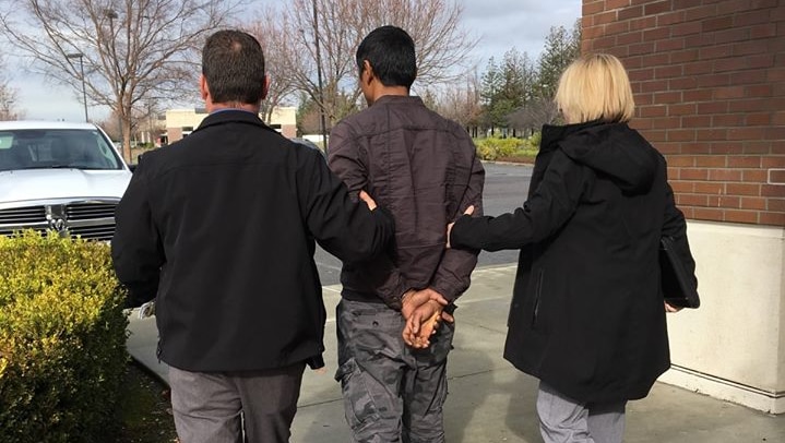 A man in handcuffs pictured from behind being led away by a male and female.