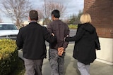 A man in handcuffs pictured from behind being led away by a male and female.