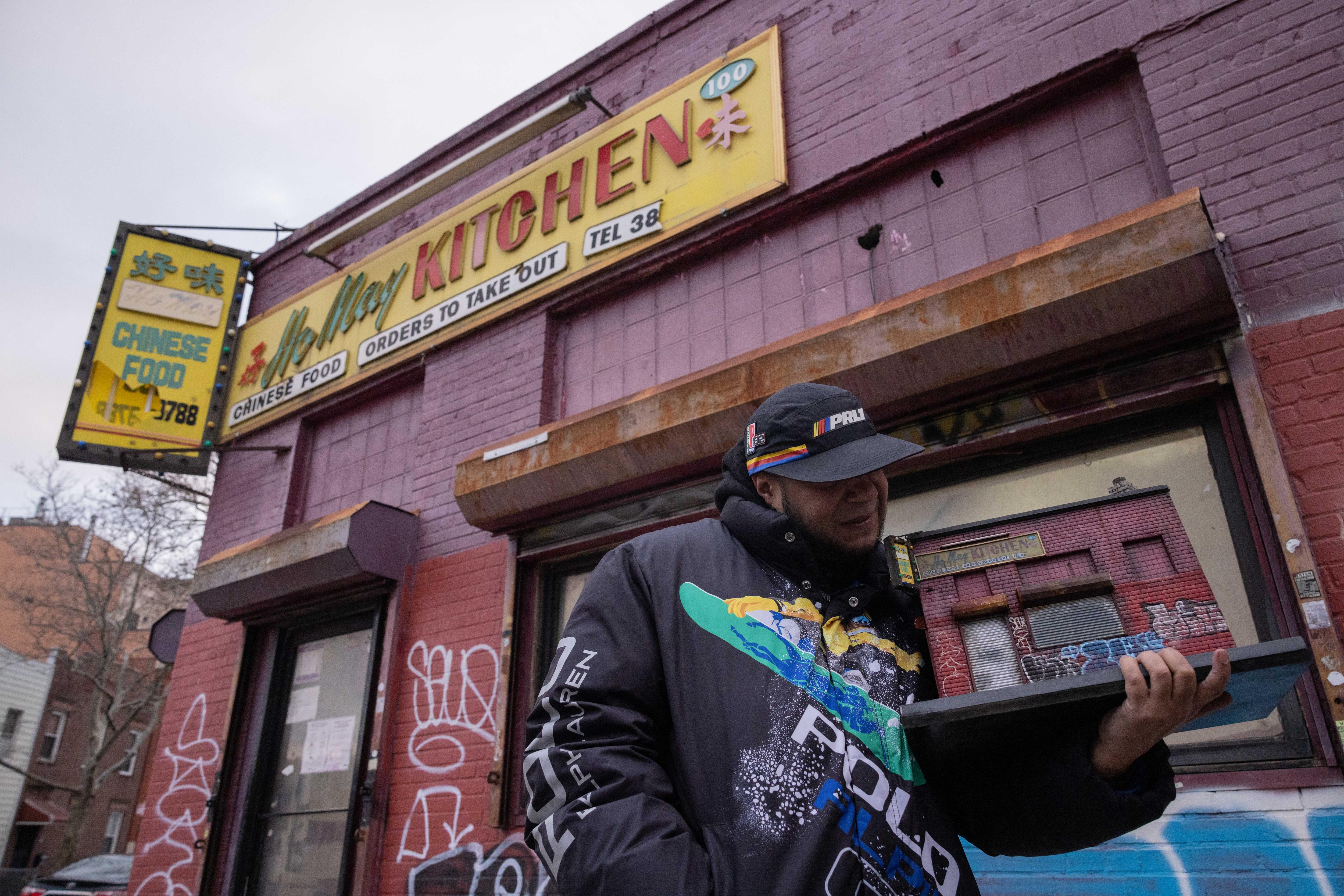 A man dressed in streetwear stands holding a miniature replica of the building behind him