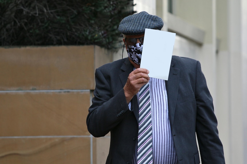 An old man wears a hat and a mask and holds a book up to hide his face 