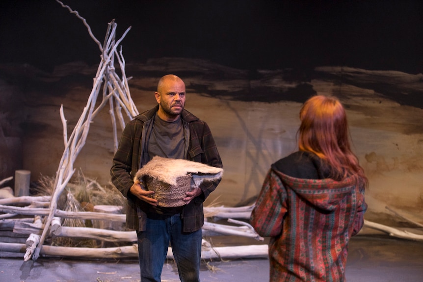 A man holding a basket and fur talks to a woman, whose face cannot be seen. Behind the man is a tipi structure made from sticks.