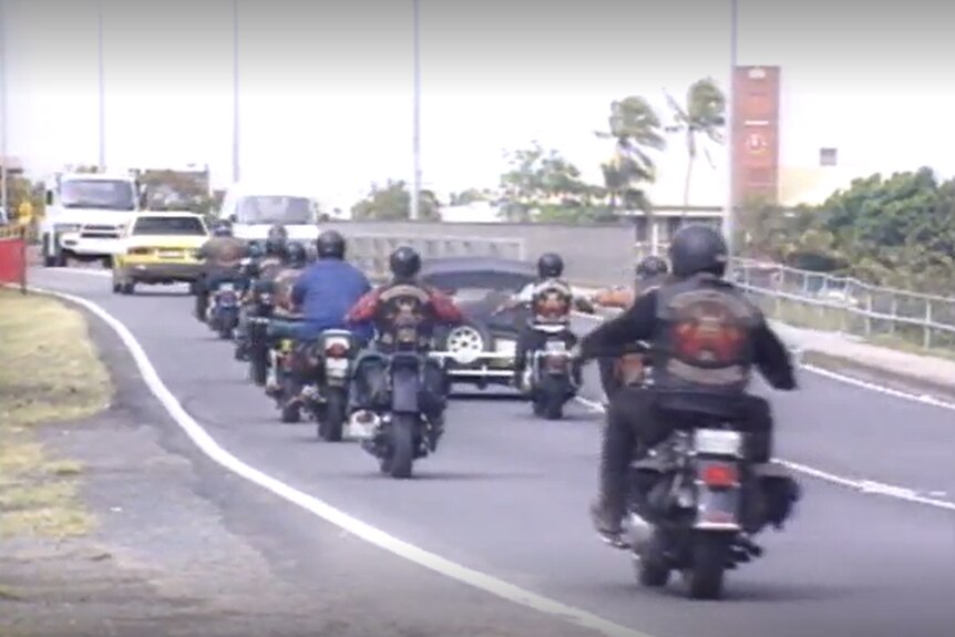 A group of outlaw motorbike riders in traffic approaching a bridge.