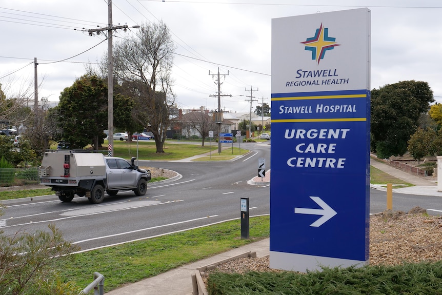 A blue and white sign with Urgent care centre with a ute on a road approaching a roundabout in the background.