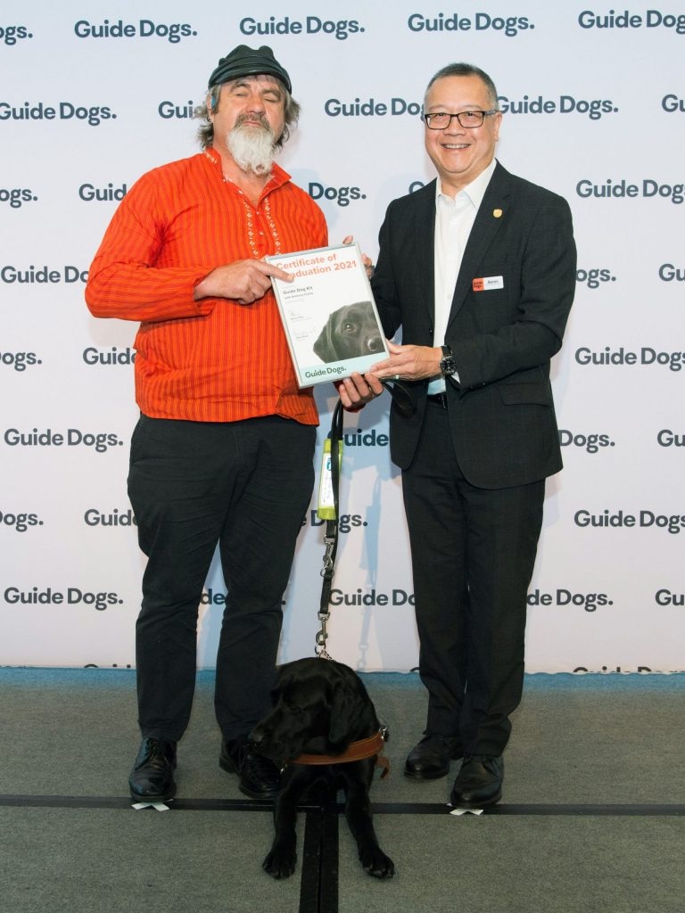 Two men holding a certificate while a black puppy sits at their feet 