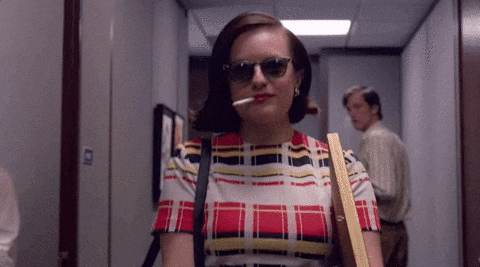 Mad Men's Peggy Olson, played by Elisabeth Moss, walks down a hallway carrying a box while smoking a cigarette.
