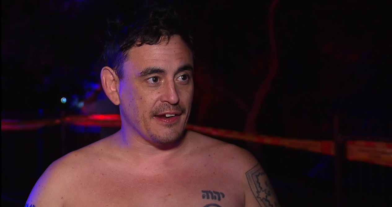 Adam Telfer tem cabelo castanho curto e tatuagens nos braços e no peito sem camisa, e está conversando com alguém.