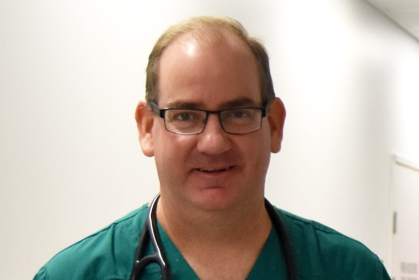 A man standing in a corridor wears hospital scrubs and a stethoscope around his neck.