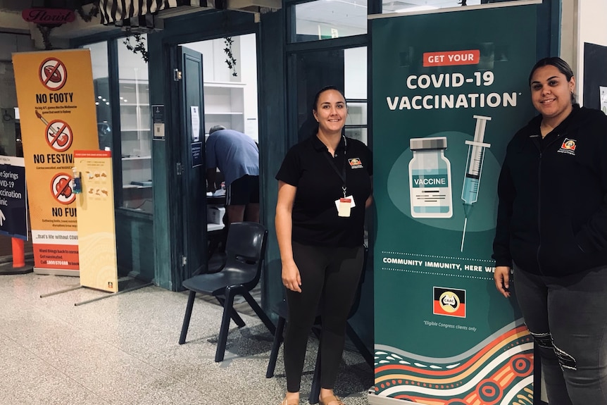 two women smile next to a 'COVID-19 vaccination' sign outside a pop up clinic