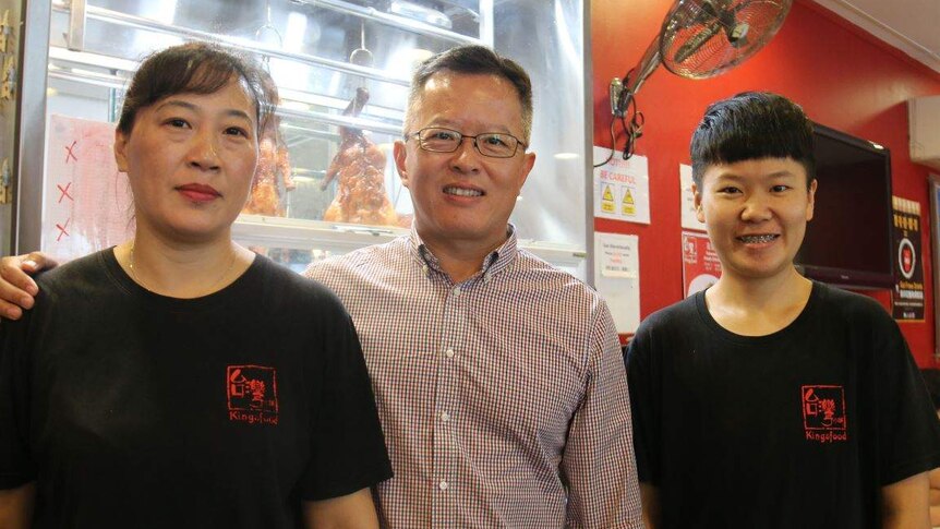 A wman and teenage boy wearing black Kingsfood shirts stand on either side of a man in a business shirt in a restaurant
