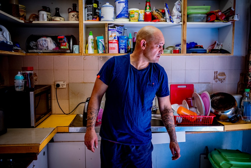 Man standing in front of a kitchen sink, looking outside. The shelves and benches are messy.