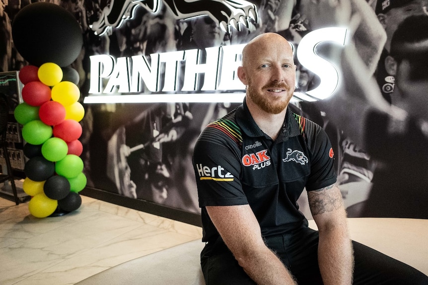 A man wearing a Penrith Panthers jersey stands in front of a Panther's sign.
