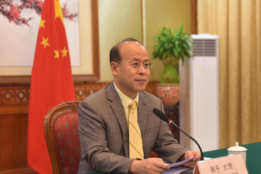 Xiao Qian sits at a desk in front of a Chinese flag.
