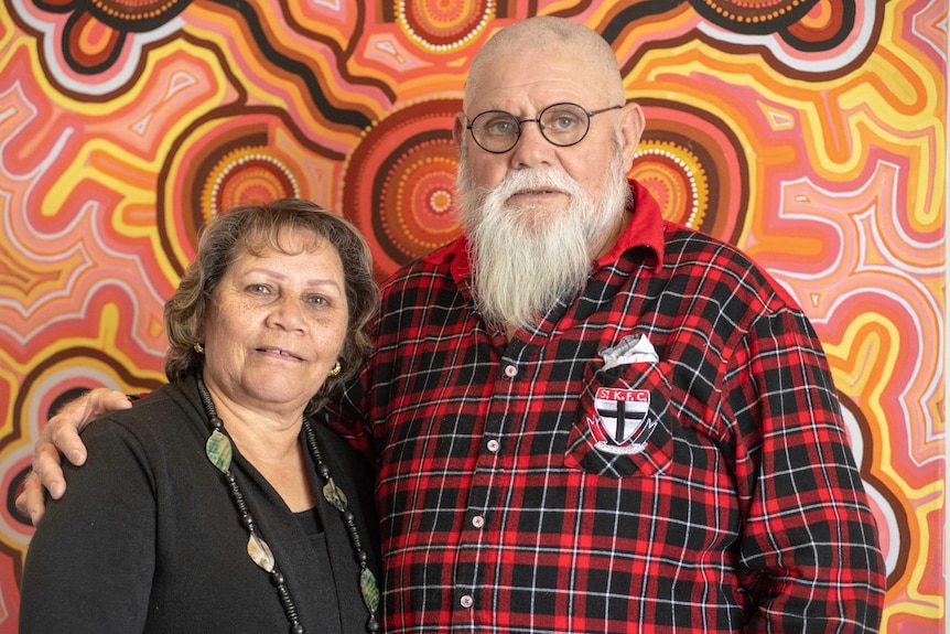 A man and a woman stand in front of an Aboriginal artwork and look at the camera.