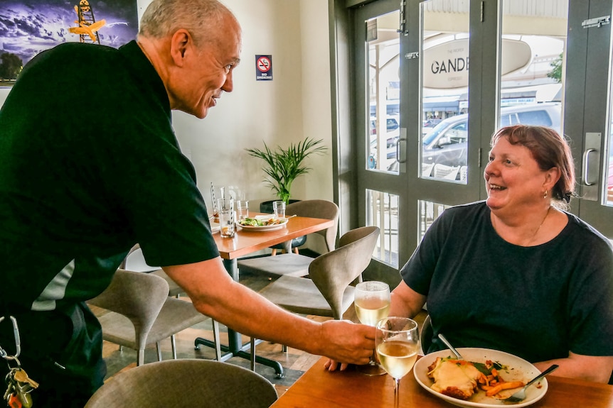 A male hospitality worker serves a woman a glass of wine with her meal in a restaurant.  