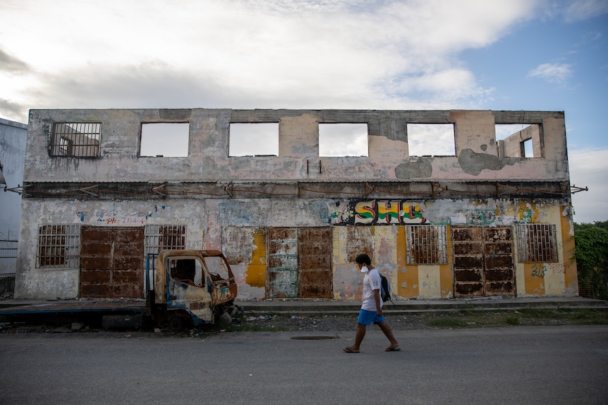 A man walks down a street past the boarded up shell of a two story building, and a burnt-out truck.