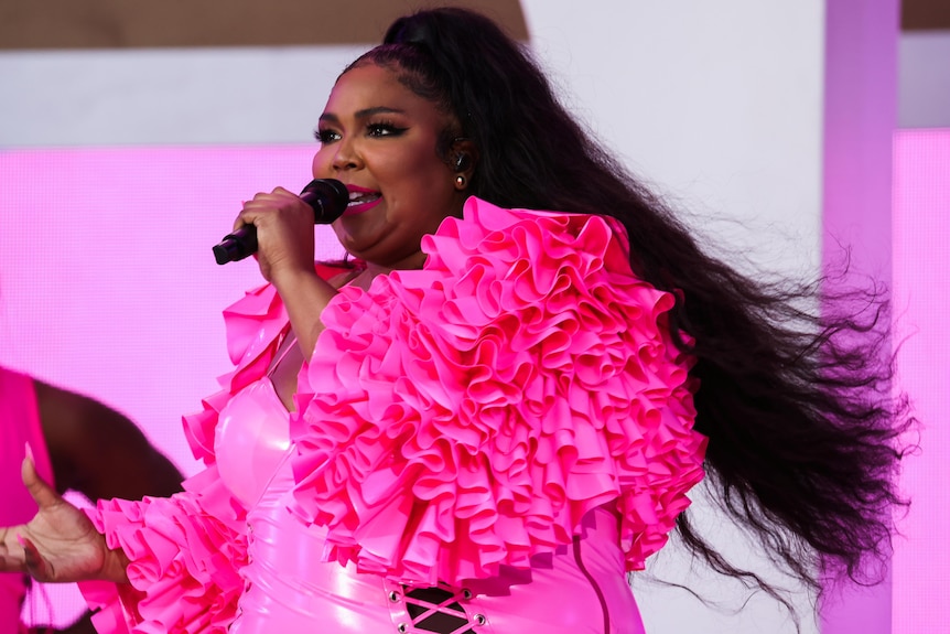 Lizzo in a pink outfit and her tied back sings at a microphone on stage in New York