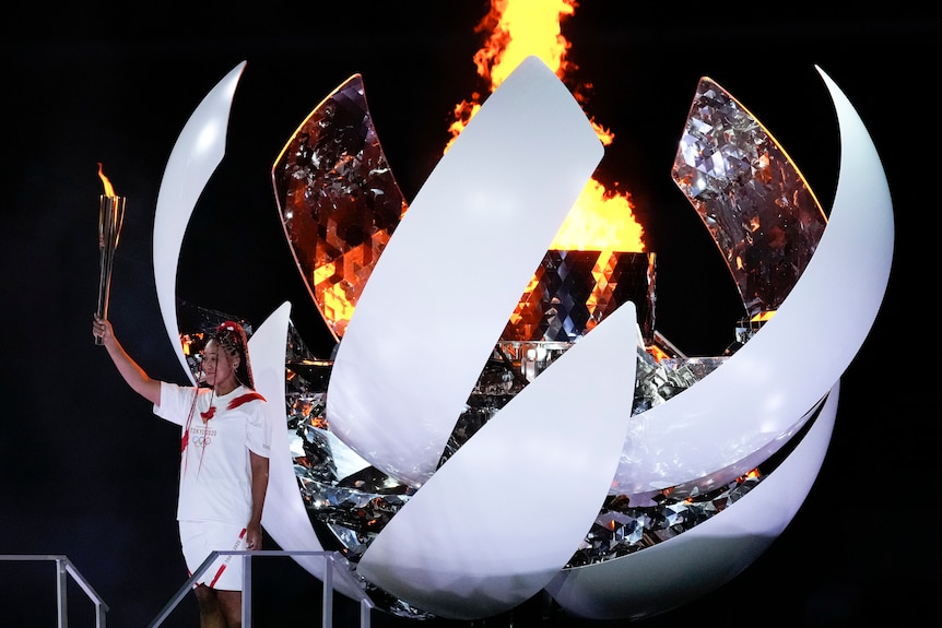 A smiling Naomi Osaka stands with the torch raised as the flame burns behind her in the Olympic cauldron. 