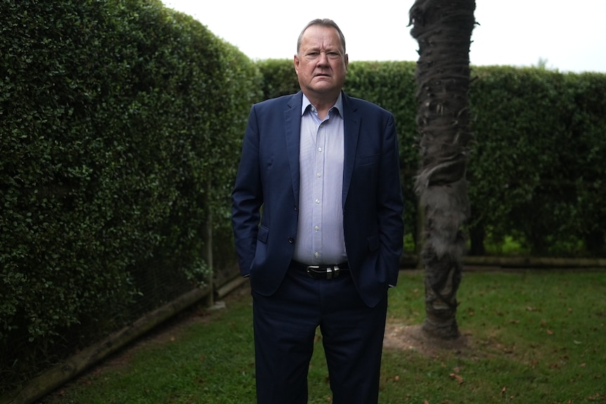 Patrick Castles stands in a yard. Behind him is a large hedge and the trunk of a tree. He has his hands in his pockets.