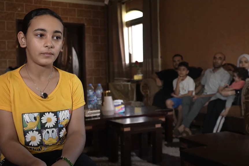 Young girl in a yellow shirt sitting in a living room with family members behind her.