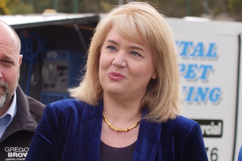 A woman with blonde hair in business attire speaks to someone off camera 