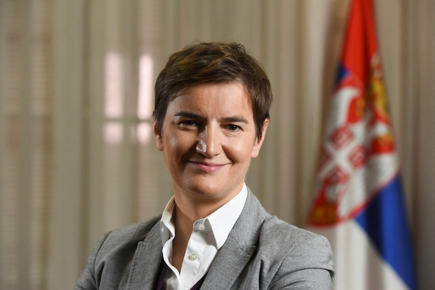 A woman in a grey suit coat and white shirt smiles as she poses for a photo in an office with a Serbian flag.