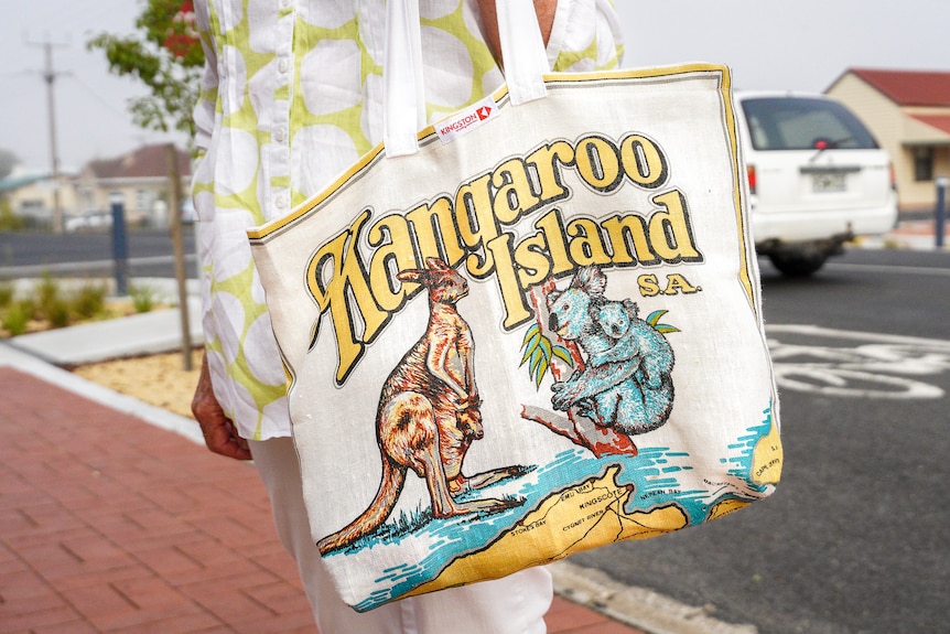 A lady standing in a street holds a white canvas bag with a retro print on it saying 'Kangaroo Island S.A'.
