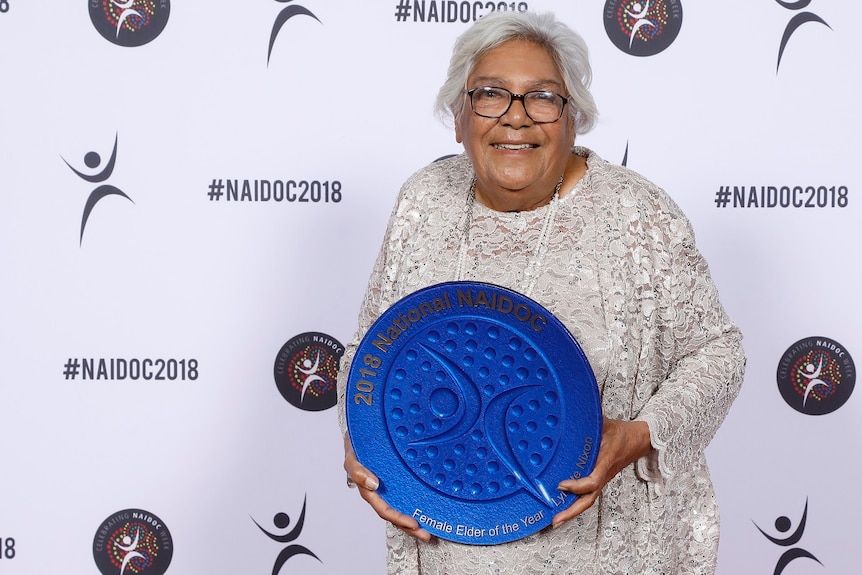 Aunty Lynette Nixon wears a silver white lace dress and black glasses as she holds a large, round, blue award. She is smiling.