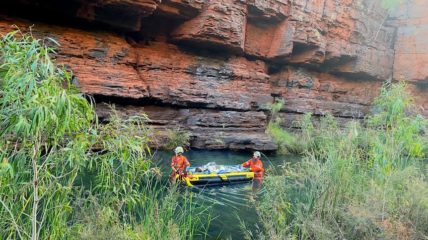 Two emergency service workers carry a woman on a stretcher through a body of water in a gorge.