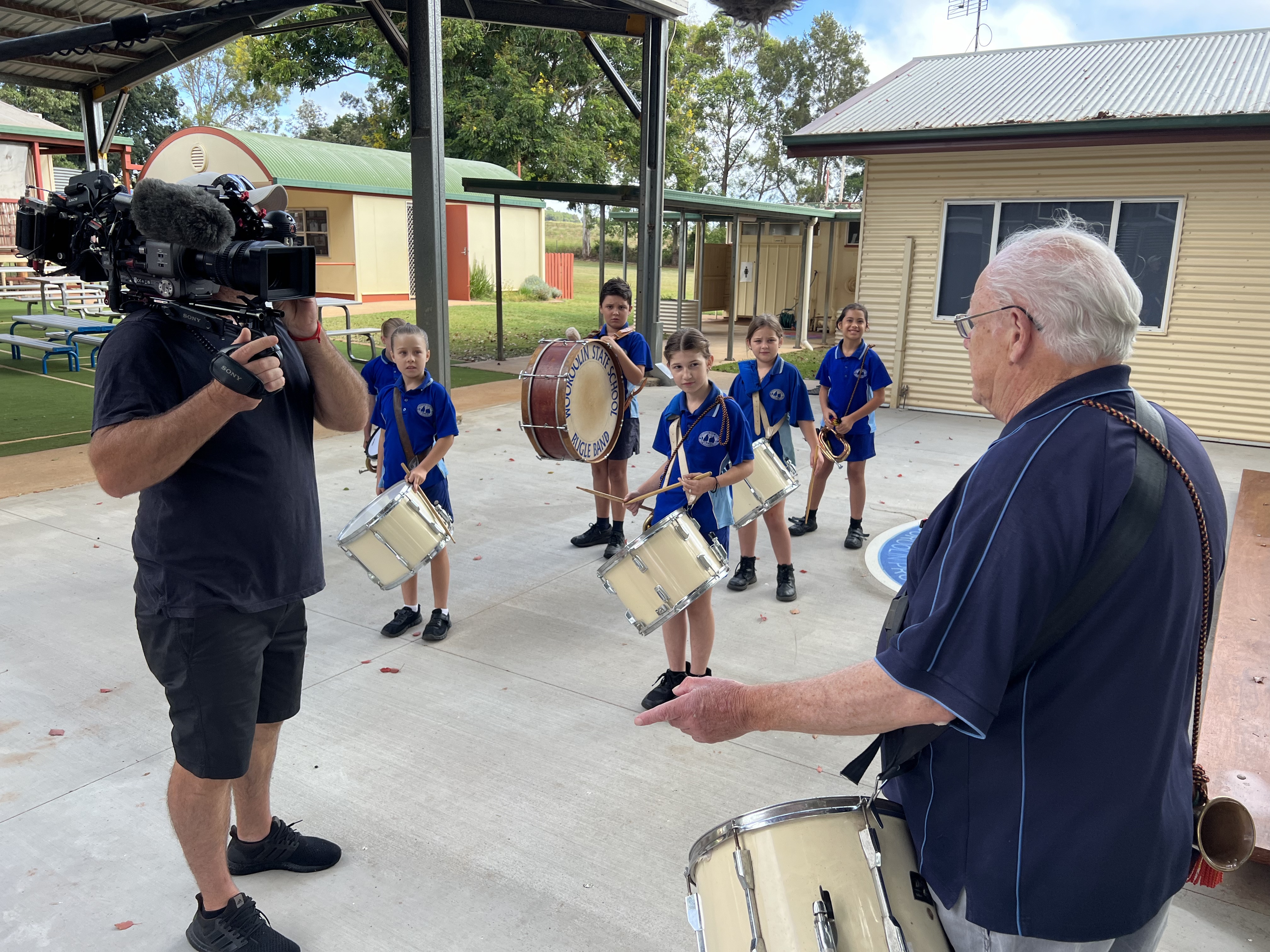 Man with back to camera speaks to a news camera in shot. Band of primary-school aged children in the background.