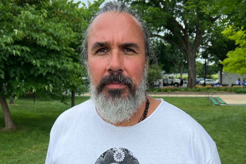 a man with a grey beard looks toward the camera with trees and grass in the background