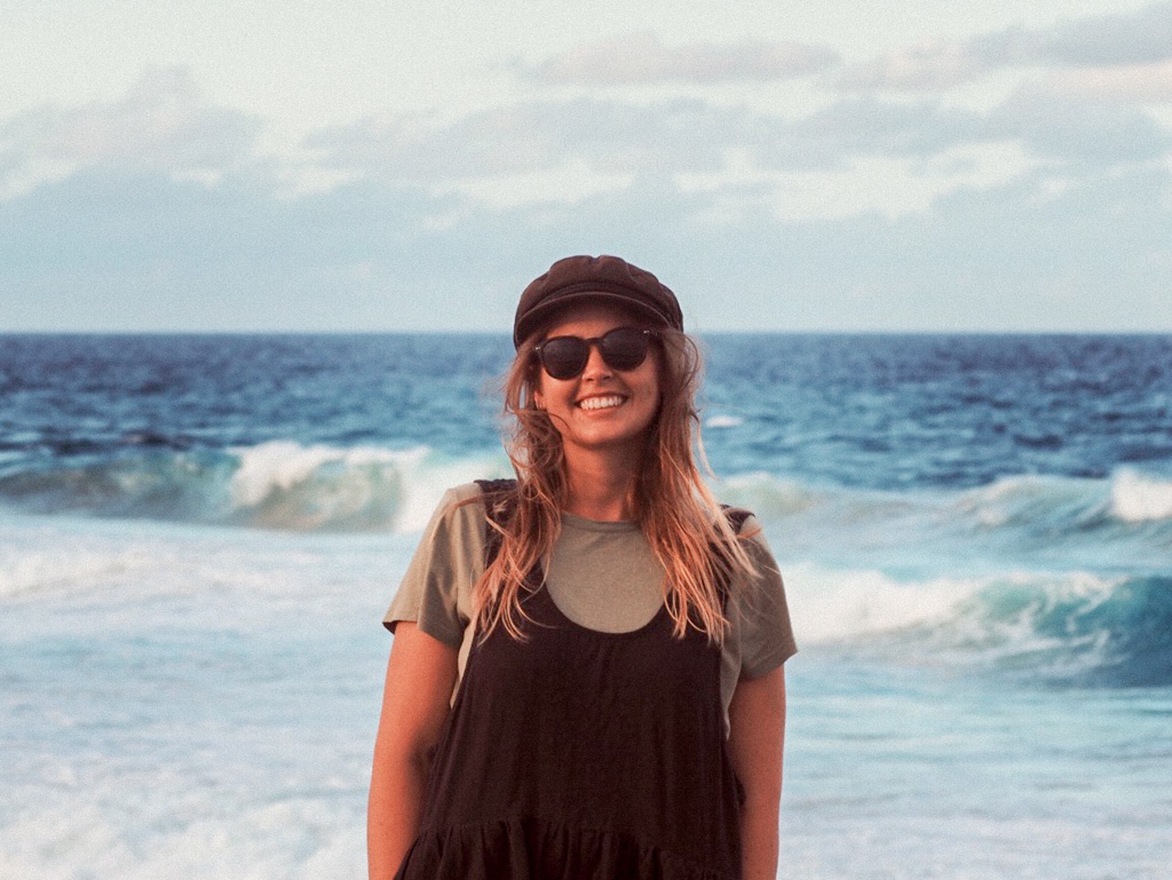 A blonde young woman wearing sunglasses and smiling stands on the beach