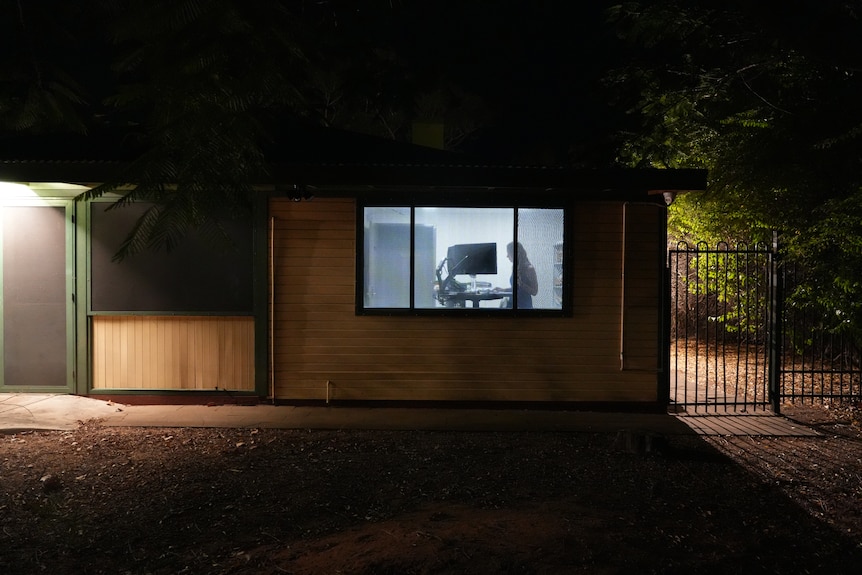 The exterior of a small office building at night with a woman on her computer behind the window.