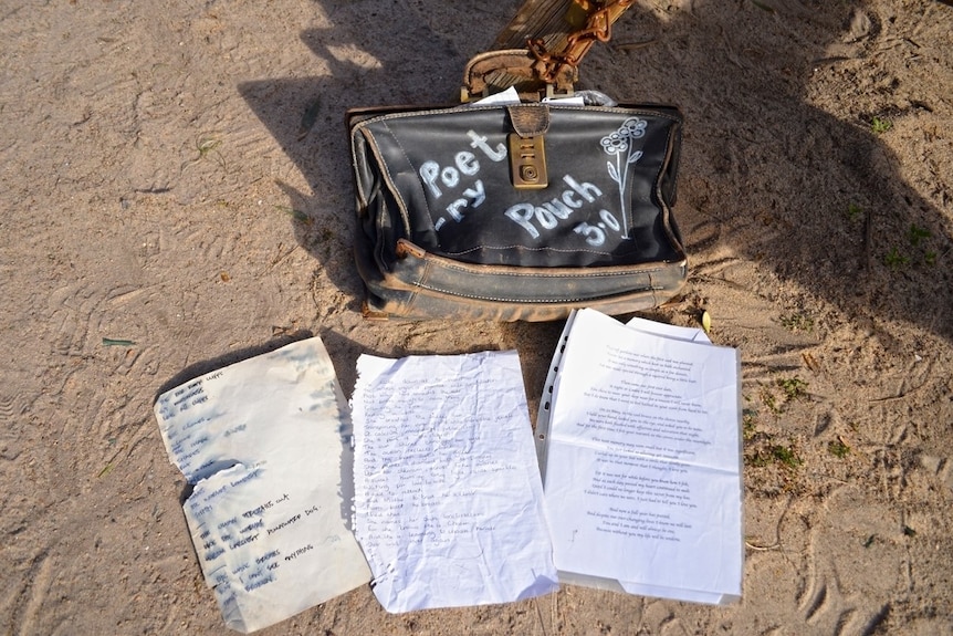 A photo of a briefcase with the words 'poetry pouch' with pieces of paper with poetry written strewn on the sand