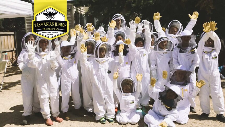 A group of children in white beekeeper suits raising their yellow gloved hands in the air.