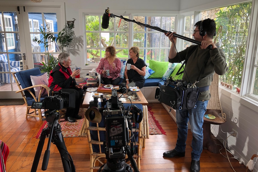 Sound recordist holding a microphone on a boom pole as three women sitting on a couch chat.