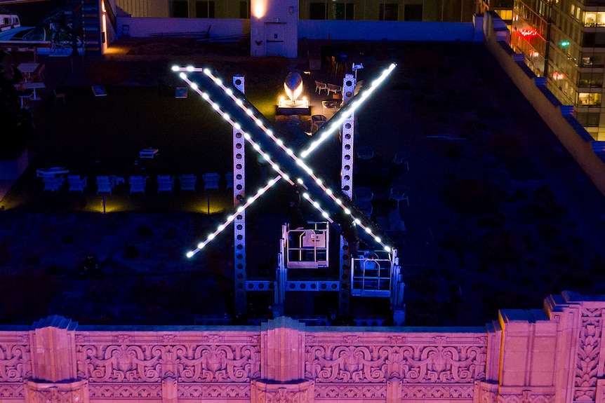 Workers installing a lit-up X logo on top of the company's headquarters building in the evening.